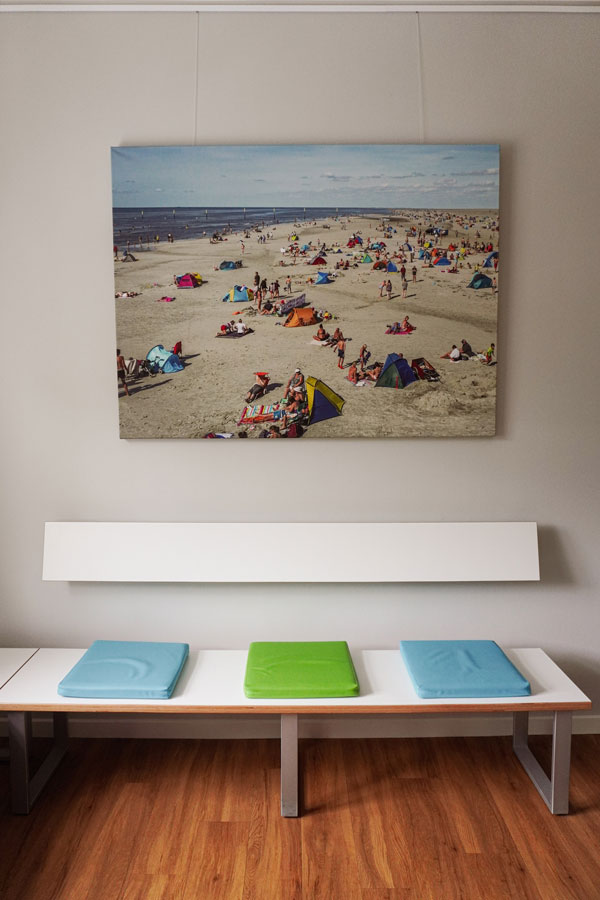 Wartezimmer mit blauen und grünen Sitzkissen, einem Holzfarbenen Bodenbelag und einer großen Fotografie von einem sommerlichen Strand mit vielen Menschen